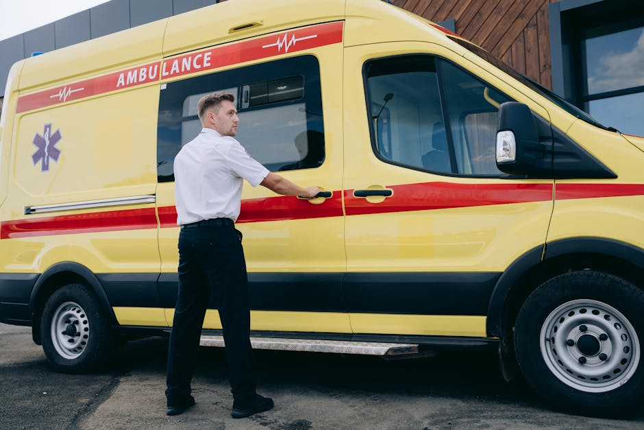 A male paramedic wearing a white uniform with dark trousers is preparing to open the sliding side door of a yellow ambulance with red and purple emergency markings. The vehicle is parked on a paved surface outside a modern building with wooden paneling and glass windows. The ambulance's design includes a large side window, a side mirror, and a black wheel with a silver rim. The background features a partly cloudy sky, and the scene appears to be set during daylight hours, emphasizing the emergency vehicle's details and the paramedic's readiness for ambulance duty.