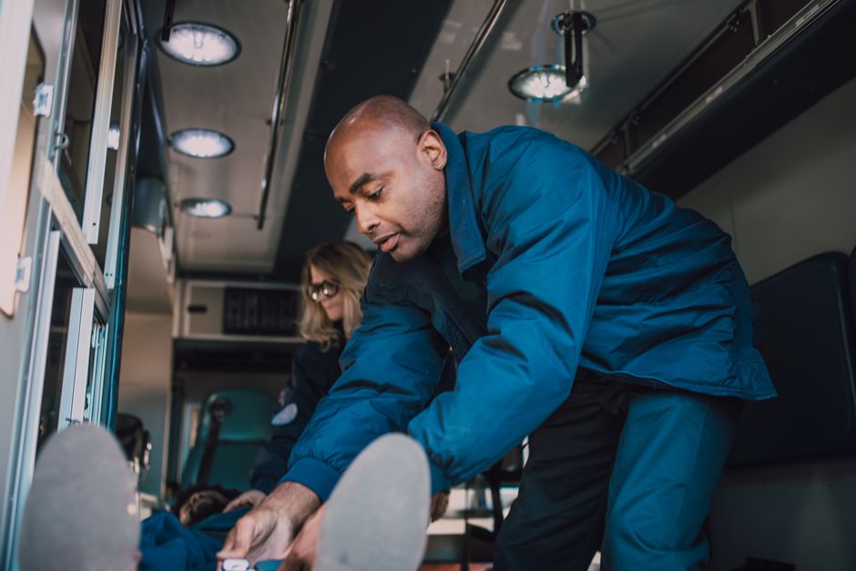 A male emergency medical technician wearing a blue uniform is inside a medical vehicle, bending forward to assist a patient by supporting their legs. Behind him, a female colleague with glasses and long hair is seated, observing the situation. The interior of the vehicle features medical equipment and storage compartments, with overhead lighting casting a bright, clinical light. The scene depicts a responsive, professional environment focused on patient care and emergency assistance.